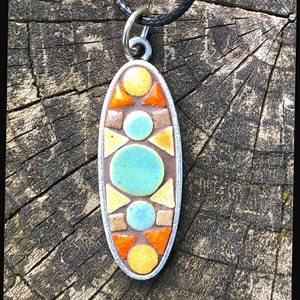 K&ouml;nnte beinhalten: Eine silberne ovale Anh&auml;nger-Halskette mit einem geometrischen Mosaik-Design. Das Mosaik zeigt orange, gelbe, braune und blaue Fliesen.