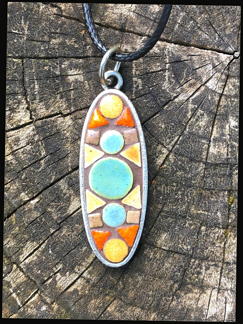 K&ouml;nnte beinhalten: Eine silberne ovale Anh&auml;nger-Halskette mit einem geometrischen Mosaik-Design. Das Mosaik zeigt orange, gelbe, braune und blaue Fliesen.