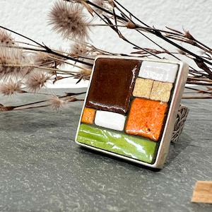 Op de afbeelding: Een vierkante ring met een mozaïekontwerp. Het mozaïek heeft bruine, oranje, witte, gele en groene tegels in een zilveren band. De ring wordt gepresenteerd op een grijze ondergrond met gedroogde planten op de achtergrond.