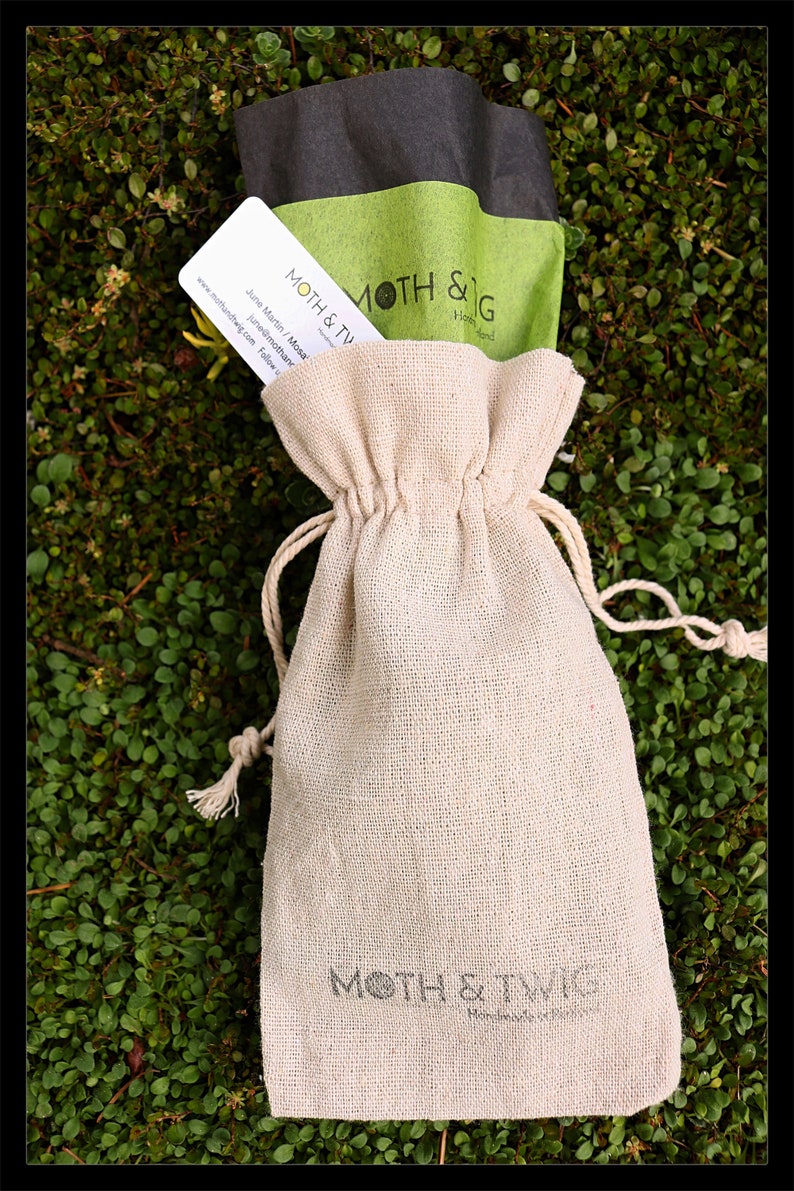 K&ouml;nnte beinhalten: Eine kleine, cremefarbene, Kordelzugtasche mit dem Aufdruck "MOTH & TWIG Handmade in Ireland" auf der Vorderseite. Die Tasche liegt auf einem Bett aus gr&uuml;nem Moos.