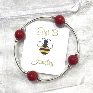May include: A silver bangle bracelet with four red beads. The bracelet is on a white background with a small white card that says "Sisi B Jewelry" with a bee logo.