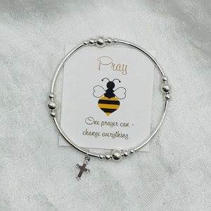 Könnte beinhalten: Ein silbernes Armband mit einem Kreuzanhänger und Perlen. Eine Karte hinter dem Armband zeigt eine Bienenillustration und die Worte "Pray" und "One prayer can change everything."