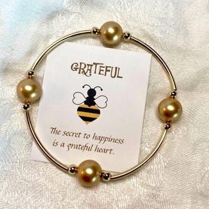 May include: A gold-colored bangle bracelet with five large, round, golden pearls. A small card inside the bracelet reads "GRATEFUL" with a bee illustration and the text "The secret to happiness is a grateful heart."