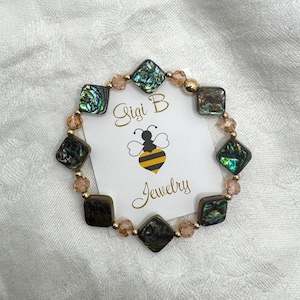 May include: A beaded bracelet featuring square abalone shell tiles with iridescent green and blue tones, interspersed with small peach-colored faceted beads and gold-tone spacers, displayed on a white card labeled Gigi B Jewelry with a bee icon.