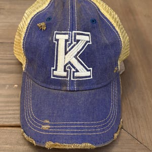 May include: A blue and yellow distressed trucker hat with a white letter K on the front.