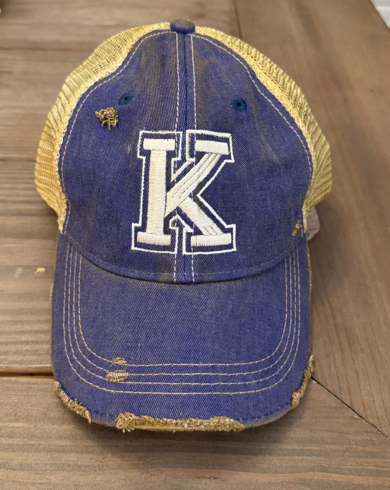 May include: A blue and yellow distressed trucker hat with a white letter K on the front.