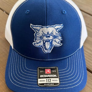 May include: A blue and white Richardson 112 Trucker hat with a wildcat logo. The hat features a blue front panel with a white embroidered wildcat and a white mesh back. The brim is blue with white stitching. The hat is adjustable.