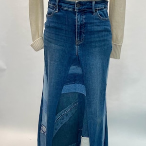 May include: A long denim skirt with a patchwork design. The skirt is made from various shades of blue denim, with different panels sewn together. The skirt has a high waist and a long, flowing silhouette. The skirt is paired with a cream-colored sweater.