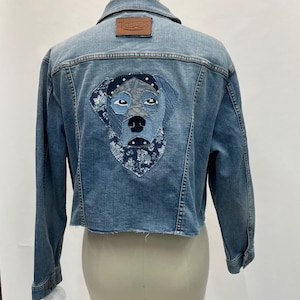 May include: Light blue denim jacket with a raw hem. The back features a patchwork dog design with a floral bandana. A brown leather patch with the text "TatterSons" is on the upper back.