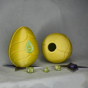 May include: Two yellow, 3D printed, egg-shaped objects with a green design. One egg has a hole in the side. Four green dice with white numbers are scattered on a white surface.