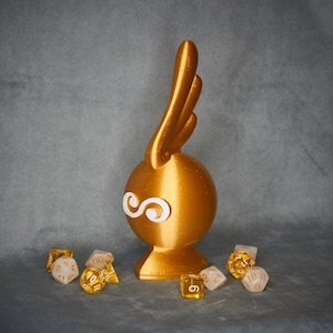 May include: A gold-colored figurine with a white wing design and a round base. The figurine is surrounded by six small, multi-colored dice.