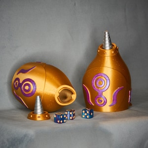 Op de afbeelding: Goudkleurige, decoratieve container met paarse accenten en een zilveren top. De container bestaat uit twee delen, met een bijpassende basis en drie blauwe dobbelstenen met gouden stippen. Steampunk- of fantasiethema.