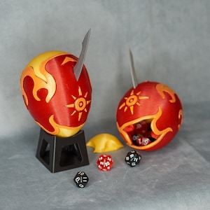 May include: Two red and yellow decorative egg-shaped containers with a sunburst design and a silver blade. One container is open, revealing dice inside. The containers are on a gray surface with dice.