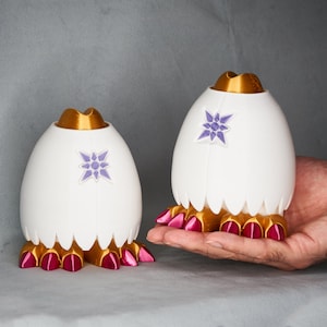 May include: Two white ceramic egg-shaped containers with gold tops and pink feet. Each container has a purple star design.
