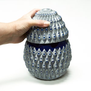 Könnte beinhalten: Ein silberfarbener dekorativer Behälter mit einer strukturierten, gemusterten Außenseite und einem blauen Innenraum. Der eiförmige Behälter hat einen Deckel und ist mit kleinen blauen Akzenten verziert. Der Behälter wird von einer Hand gehalten.