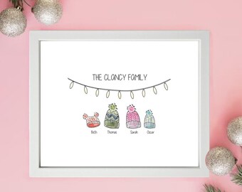 May include: A framed print featuring the text "THE CLANCY FAMILY" and illustrations of four winter hats. Each hat is a different colour and style, with names underneath. The background is white, and the frame is white.
