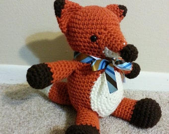 May include: A crocheted fox plush toy with brown and orange fur, white belly, and a blue and white striped ribbon around its neck.