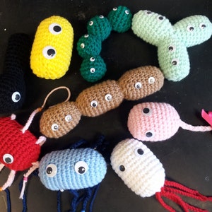 May include: A collection of crocheted plush toys shaped like different types of bacteria. The toys are in various colors, including green, yellow, brown, pink, red, and blue. Each toy has googly eyes and a crocheted texture.