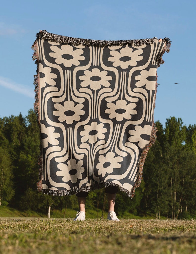 May include: A black and beige woven throw blanket with a repeating pattern of stylized flowers. The blanket has fringe on all sides.