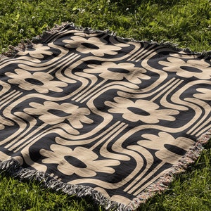 May include: A black and beige woven throw blanket with a geometric pattern of stylized flowers. The blanket has a fringed edge and is laid out on a grassy surface.