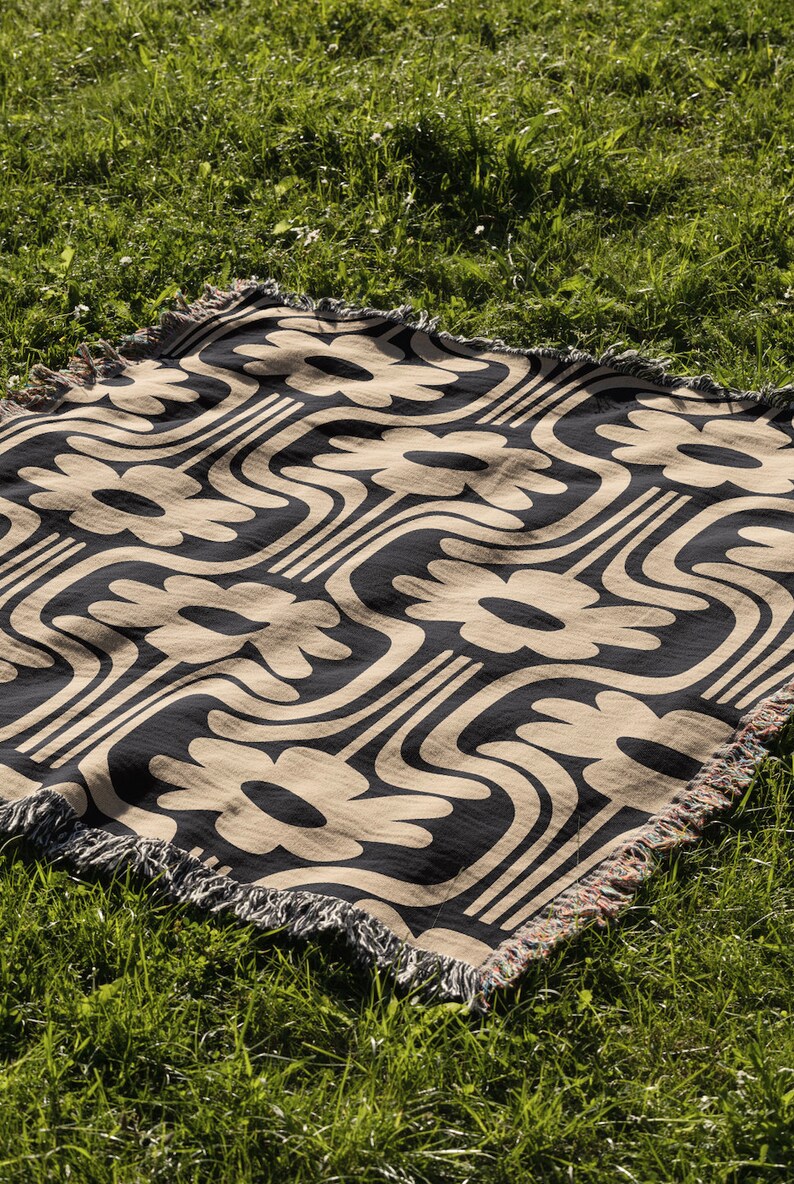 May include: A black and beige woven throw blanket with a geometric pattern of stylized flowers. The blanket has a fringed edge and is laid out on a grassy surface.
