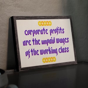 May include: A framed cross-stitch with a purple and yellow design. The text reads "corporate profits are the unpaid wages of the working class".