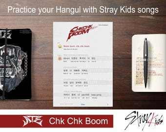 May include: A worksheet with Korean lyrics from the Stray Kids song "Chk Chk Boom". The lyrics are written in Hangul, the Korean alphabet. The worksheet is on a wooden table with a notebook and pen.