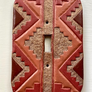 May include: A rectangular light switch cover with a geometric design. The cover is made of leather and features a pattern of triangles and stepped shapes in shades of red, brown, and tan. The center has an opening for the switch and two screw holes.