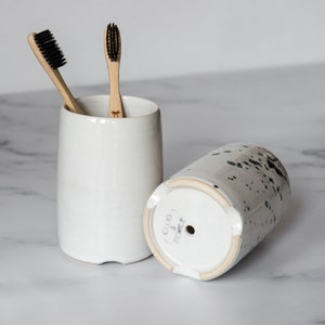 May include: Two white ceramic toothbrush holders with black speckles. One holder has the text "CLOUD & PEPPER" on the bottom. Two bamboo toothbrushes with black bristles are in the holder.