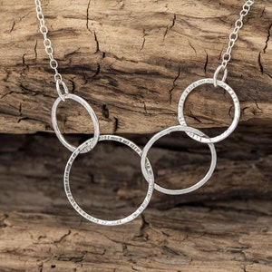 May include: A silver necklace featuring four interlinked circles, each with a textured finish. The necklace is displayed against a wooden backdrop.