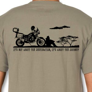 Può includere: Una t-shirt color cachi con una grafica nera di una persona che guida una moto con il testo "Non è la destinazione che conta, è il viaggio."
