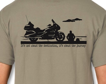 May include: A light olive green short-sleeve t-shirt featuring a black graphic on the back. The design depicts a motorcycle and a person sitting nearby, with the text "It's not about the destination, it's about the journey" printed below.