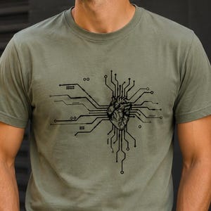 Könnte beinhalten: Ein olivgrünes Kurzarm-T-Shirt mit einem schwarzen Grafikdruck auf der Brust. Das Design zeigt ein anatomisches menschliches Herz, das in Linien und Knoten einer elektronischen Leiterplatte integriert ist, die sich horizontal erstrecken.