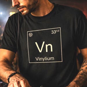 May include: A black t-shirt with a white graphic that reads "Vn Vinylium" in a periodic table style design. The DJ is wearing headphones and is in a crowded club with a neon sign that says "Vinyl Sounds Better".