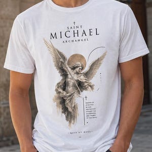 May include: A white short-sleeved t-shirt featuring a graphic print of Saint Michael the Archangel with wings and a sword. The text reads Saint Michael Archangel, Defend us in battle, be our protection against the wickedness and snares of the devil, Quis ut Deus.