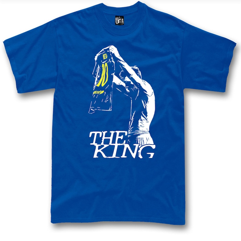 May include: Royal blue t-shirt featuring a white graphic of a person holding up a jersey with the number 10. The text "THE KING" is printed in white below the graphic. The shirt has short sleeves and a crew neck.