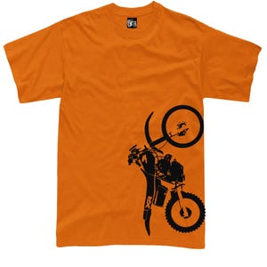 May include: Orange t-shirt featuring a black graphic of a motorcycle doing a wheelie. The motorcycle design includes details of the wheels, frame, and rider. The shirt has a crew neck and short sleeves.