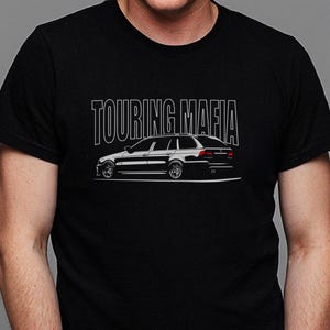 Puede incluir: Camiseta negra con la frase "TOURING MAFIA" sobre una ilustración blanca de un coche familiar. El coche se representa de perfil, mostrando detalles como las ruedas y las ventanas. La camiseta tiene cuello redondo.