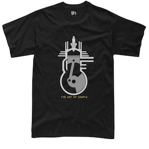Może przedstawiać: Czarny t-shirt z krótkim rękawem z szarą grafiką przedstawiającą tłok silnika spalinowego oraz świecę zapłonową. Poniżej grafiki na przodzie odzieży znajduje się nadruk z napisem THE ART OF SIMPLE wykonany czcionką w kolorze złotym.