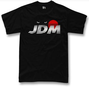 Op de afbeelding: Zwart T-shirt met het woord "JDM" in wit en zilver, een rode zon en gestileerde ogen boven de tekst. Het shirt heeft korte mouwen en een ronde hals.