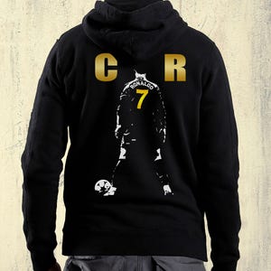 Peut inclure: Un sweat-shirt à capuche noir avec un logo CR doré sur le haut du dos. Sous le logo se trouve une silhouette graphique blanche d'un joueur de football avec le nom RONALDO et le numéro 7 imprimés en jaune au dos du maillot du joueur.