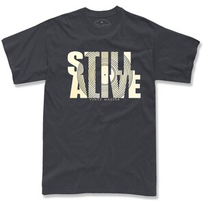 A black t-shirt with a graphic design featuring the text "STILL ALIVE" in a stylized font, with the word "VINYL" underneath.