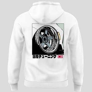 Könnte beinhalten: Weißer Kapuzenpullover mit einer Grafik eines Autorads mit Chromfelge und blauem Mittelteil. Die Grafik befindet sich in einem schwarzen Rechteck, darunter japanischer Text und ein Sonnenaufgangssymbol. Der Hoodie hat eine Kapuze mit Kordelzug.
