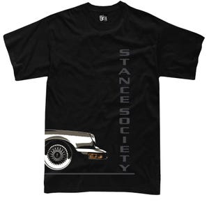 May include: Black t-shirt featuring a car's front quarter panel with a chrome wheel and the text "STANCE SOCIETY" in gray vertical lettering. The shirt has a classic crew neck and short sleeves.