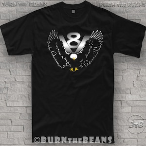 May include: Black t-shirt featuring a silver V8 logo with an eagle design. The eagle has outstretched wings and a white head with yellow feet. The text "BURN THE BEANS" is printed at the top and bottom of the shirt.