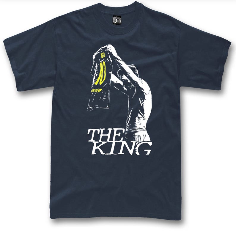 May include: Navy blue t-shirt featuring a white graphic of a person holding up a jersey with the number 10 in yellow. The text "THE KING" is printed in white below the graphic. The shirt is a classic crew neck style.