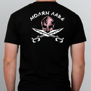 以下が含まれることがあります： 背中に白いグラフィックが入った黒いTシャツ。グラフィックは、2本の交差した剣の上にピンク色のグラディエーターヘルメットがあり、ギリシャ語のフレーズ「ΜΟΛΩΝ ΛΑΒΕ」が白い文字で書かれています。