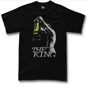 May include: Black t-shirt featuring a gray silhouette of a person holding up a jersey with the number 10 in yellow. The text "THE KING" is printed in gray below the figure. The shirt is a classic crew neck style.
