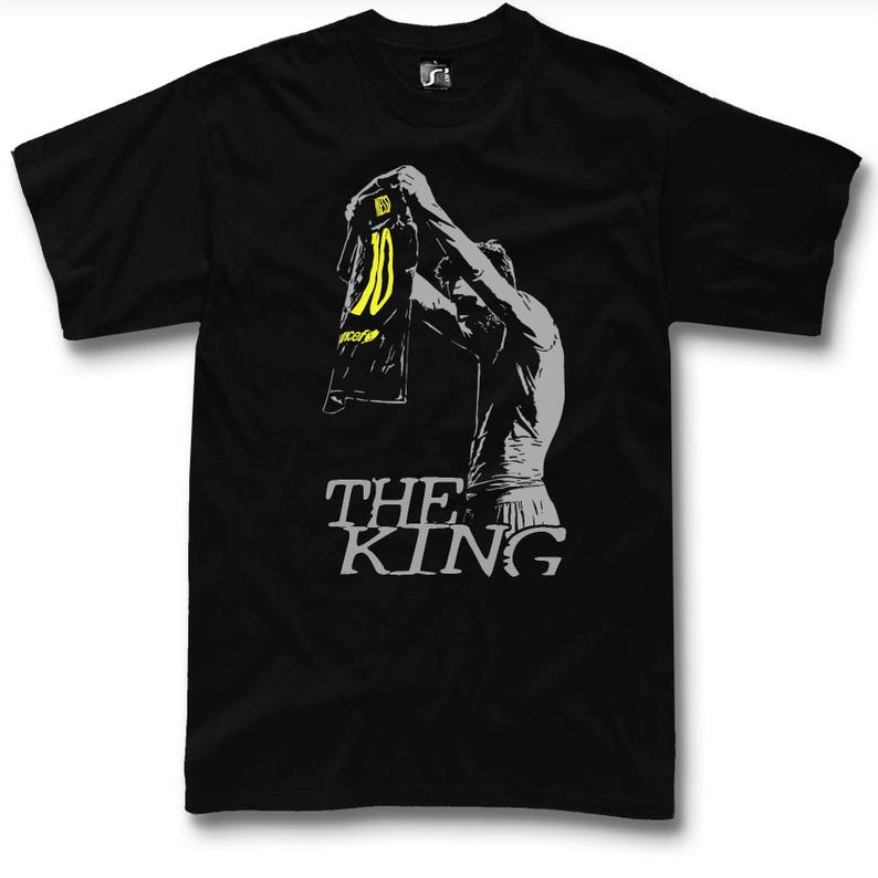May include: Black t-shirt featuring a gray silhouette of a person holding up a jersey with the number 10 in yellow. The text "THE KING" is printed in gray below the figure. The shirt is a classic crew neck style.