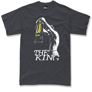 May include: Dark gray t-shirt featuring a white graphic of a person holding up a jersey with the number 10 in yellow. The text "THE KING" is printed in white below the graphic. The shirt is a crew neck.